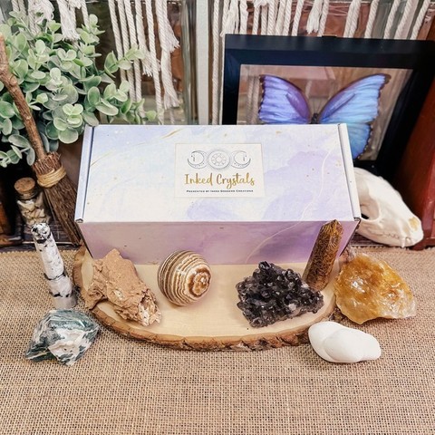 A photo of our exclusive "Inked Crystals" subscription box product. It features our packaging as well as examples of past specimens that subscribers have received. Each specimen comes with an Eclectic Witch Card Deck card and a printed book of shadows page about the mineral. Presented by Inked Goddess Creations.