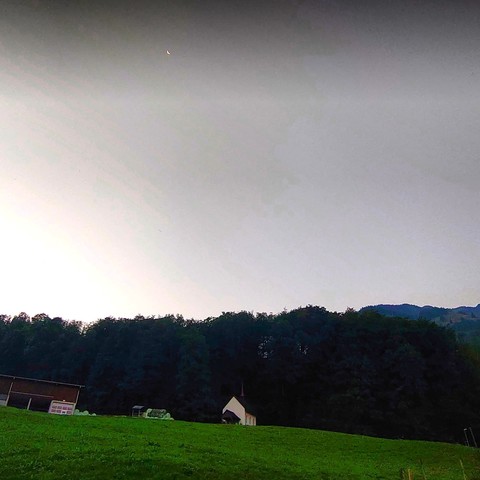 Blick in den Morgenhimmel. Unten im Bild eine Wiese, mittig die Lourdeskapelle. Dahinter Wald und Berge. Darüber der Morgenhimmel und die Mondsichel. 