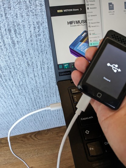 I small Mp3 player held in a hand connected to a laptop using a USB C to USB C cable transfer files and charging