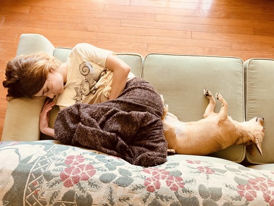 A teen girl with honey-blonde hair, a light T and brown fuzzy pants sleeps curled on her side on an ice-green couch, butt-to-butt with her beige daschund-mix therapy pup, who is also sacked out. 