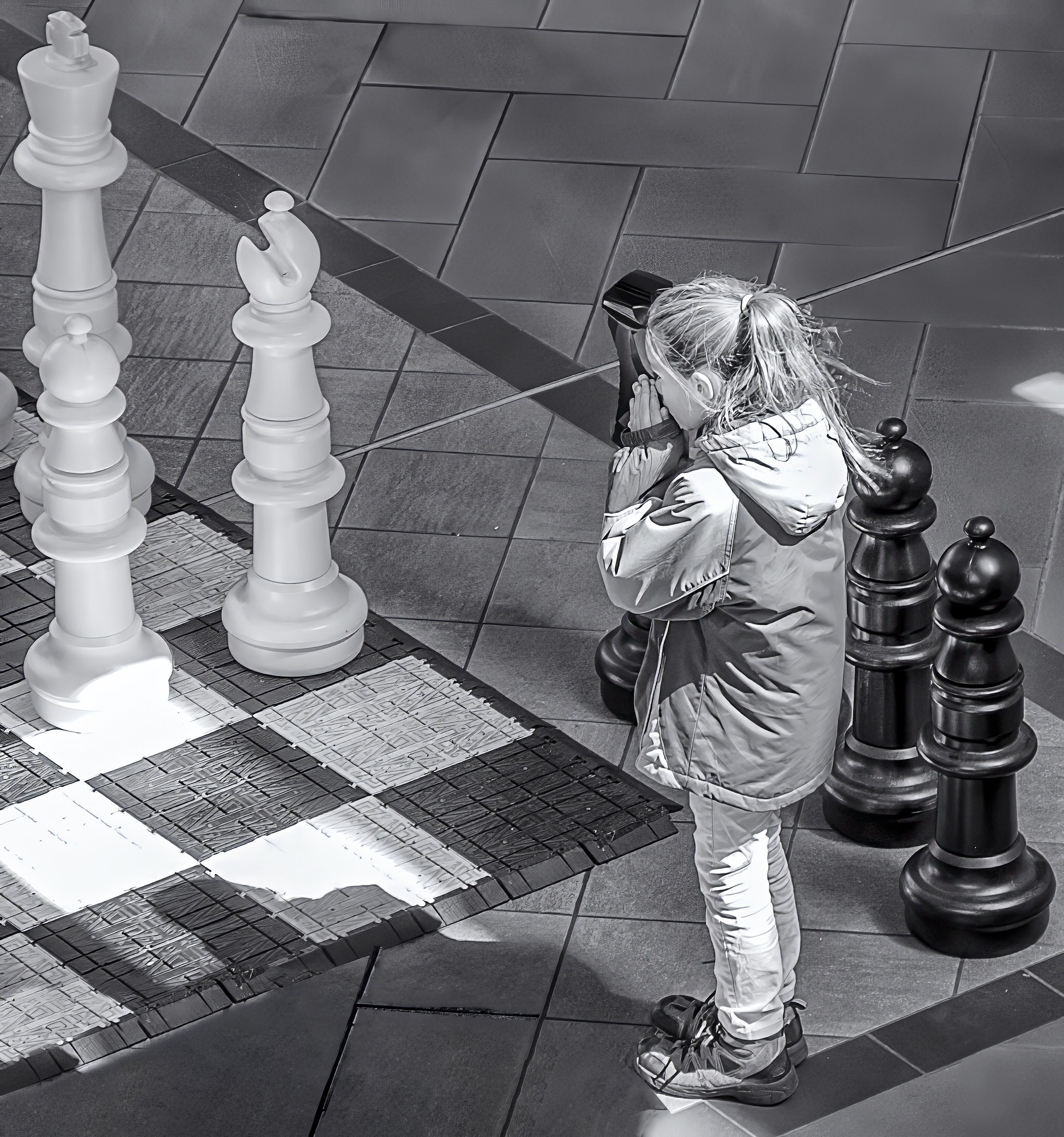 Young girl stands in front of a supersize chess board contemplating the position of the piece on the board. 