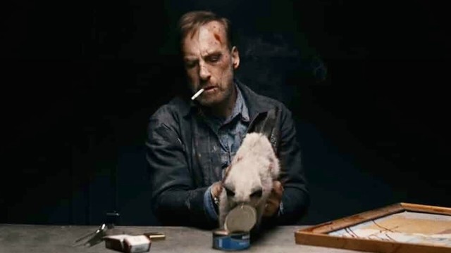 Bob Odenkirk as the titular character in NOBODY (20121). He has a cigarette in his mouth, looks very bruised and battered, and is sitting at a table helping a cat eat from an open tin of wet food 