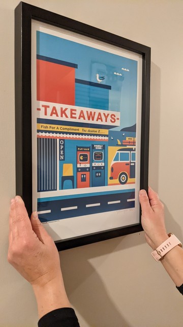 Hanging a print called Tiki Tour Takeaways by Greg Straight on the wall. Hands holding up the framed print to the wall to decide if this is the perfect spot for it.