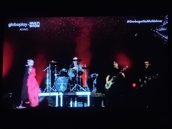 PT-BR: Foto de televisão mostrando o festival The Thown 2023 no canal a cabo Multishow. Aparece uma visão geral da banda Garbage. Da esquerda para a direita estão Shirley Manson, vocalista da banda, o bateirista Butch Vig, o baixista e o guitarrista. O fundo está preto com um jogo de luzes e fumaça com a cor vermelha. Há pontinhos de luz na parte de cima, como um céu noturno estrelado. Na tela, no canto superior esquerdo está escrito, em letras brancas: globoplay + MULTISHOW (logotipo) com AO VIVO embaixo. No canto superior direito esta escrito em letras brancas e em itálico: #GarbageNoMultishow.