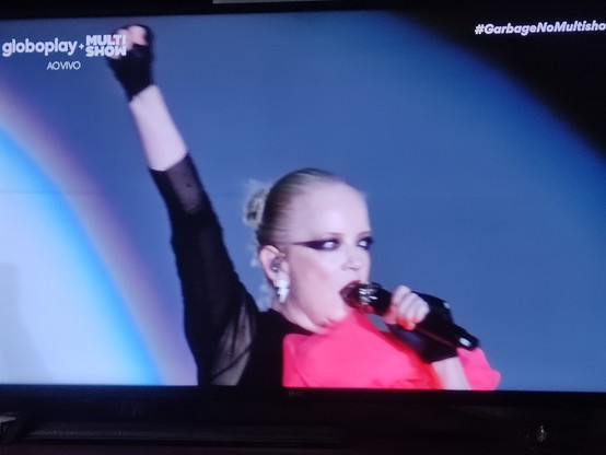 PT-BR: Foto de televisão mostrando o festival The Thown 2023 no canal a cabo Multishow. Aparece Shirley Manson, vocalista da banda Garbage, do peito para cima, cantando, segurando o microfone com a mão esquerda e erguendo o braço direito para cima com o punho cerrado. Ela está com o cabelo preso, maquiagem preta em volta dos olhos se estendendo até as orelhas quase como uma máscara. Usa uma roupa preta semitransparente com a manga indo até o meio do antebraço e um tecido rosa volumoso costurado na frente. Nas mãos ela usa luvas meio dedo pretas. No fundo predomina a cor azul sem objetos, apenas reflexo de luzes. Na tela, no canto superior esquerdo está escrito, em letras brancas: globoplay + MULTISHOW (logotipo) com AO VIVO embaixo. No canto superior direito esta escrito em letras brancas e em itálico: #GarbageNoMultishow.