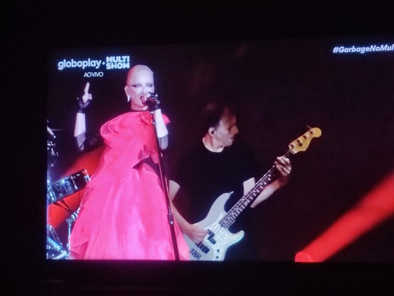 PT-BR: Foto de televisão mostrando o festival The Thown 2023 no canal a cabo Multishow. Aparecem dois integrantes da banda Garbage. À esquerda está Shirley Manson cantando, com o corpo virado para a esquerda e a cabeça para frente. Ela segura o microfone com a mão esquerda e está com a mão direita, na altura da cabeça, com o dedo em riste apontado para cima. Atrás dela e à direita está o baixista, homem branco vestindo camisa preta. O fundo está quase todo preto com uma faixa de luz vermelha na parte inferior direita da imagem. Na tela, no canto superior esquerdo está escrito, em letras brancas: globoplay + MULTISHOW (logotipo) com AO VIVO embaixo. No canto superior direito esta escrito em letras brancas e em itálico: #GarbageNoMultishow.