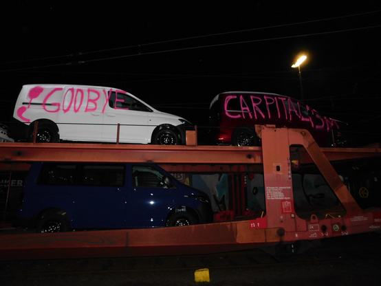 Ein Autozug auf dem mehrere Autos stehen bei Nacht. Auf den Autos in der oberen Reihe steht mit pinker Farbe geschrieben: “ Good Bye Carpitalism”