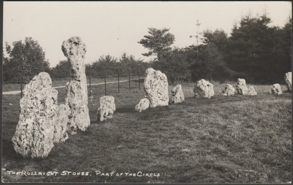 A vintage black and white photographic postcard showing an arc of the King's Men stone circle at the Rollright Stones in Oxfordshire, close to the border with Warwickshire. A dolmen called the Whispering Knights is on the opposite side of the same field and an isolated menhir, the King Stone, stands across the county border on the other side of the road. The stones of all three are oolitic limestone. Frank Packer is known to have reused some of the photos originally taken by Percy Simms, also of Chipping Norton, a couple of decades earlier.