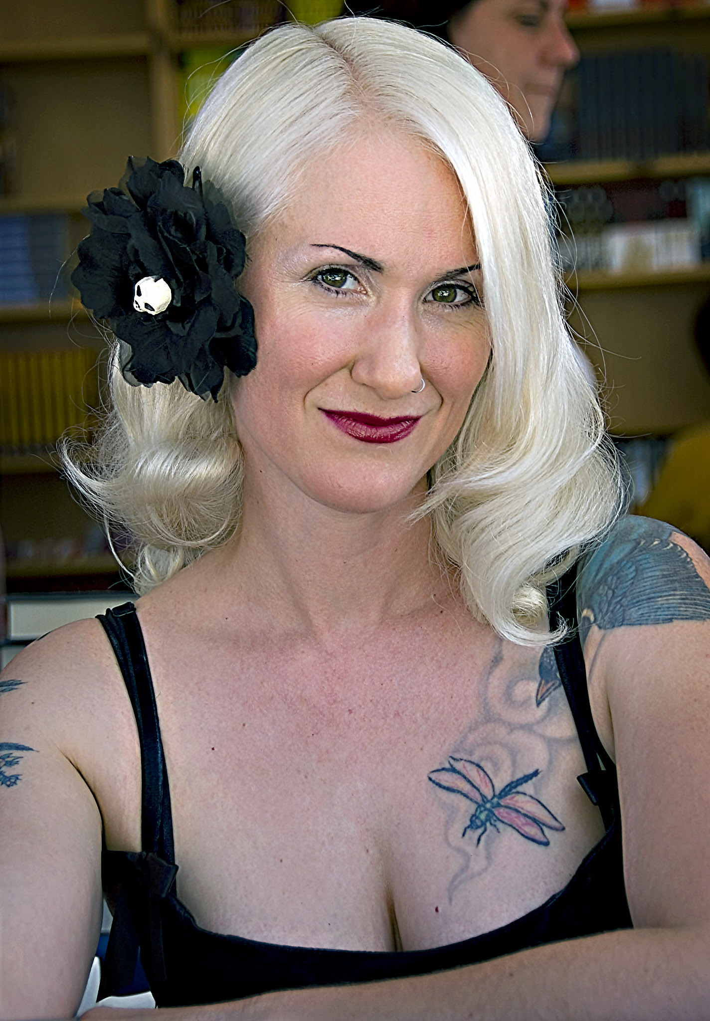 Author Christa Faust signing at a booth at the LA Times Festival of Books. She has a black hair barrette that looks like a flower with a tiny skull in the middle and she is wearing a black strappy dress that shows off several of her tattoos.