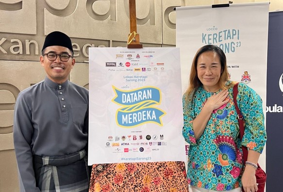Two person with the signboard that revealed the location for Keretapi Sarong 2023 event to be at Dataran Merdeka. Photo is from the article linked.