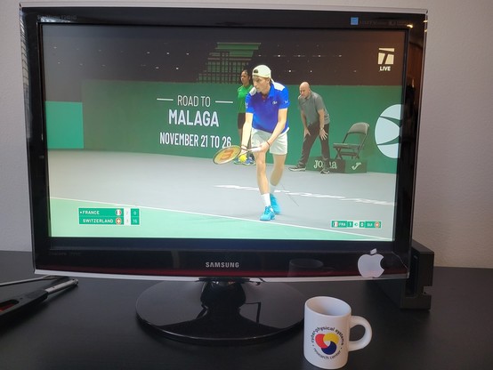 A Samsung television showing the broadcast of the 2023 Davis Cup, Group B tennis tournament on day 1. The screen shows French male tennis player Ugo Humbert serving on AO Arena. Humbert wears a blue & white Lacoste top with a reversed white cap, white shorts, white athletic socks, and blue shoes, with a Wilson tennis racquet. The wall behind him reads Road to Malaga November 21 to 26. The court color is traditional green and is surrounded by a light gray surface. In the bottom-left is the score, with France  (Humbert) leading Switzerland (Wawrinka) in the 1st set 2-1 at 0-15 in the current game.

In front of the TV is a white, 3 ounce, ceramic espresso (demitasse) cup with a shot of espresso, on a dark brown-black media console table. The blue text on the cup reads "Cyber-Physical Systems Research Center" and wraps in a circle around the logo, which looks like a loosely interlaced, rounded triquetra with thick knots in solid red, yellow, and blue (each its own color).