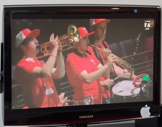 Four-member band in the stands: 1) Trumpet; 2) Trumpet; 3) Soprano saxophone; and marching-band snare drum/tambourine combination. They wear matching red shirts and hats emblazoned with Swiss flag symbols. 