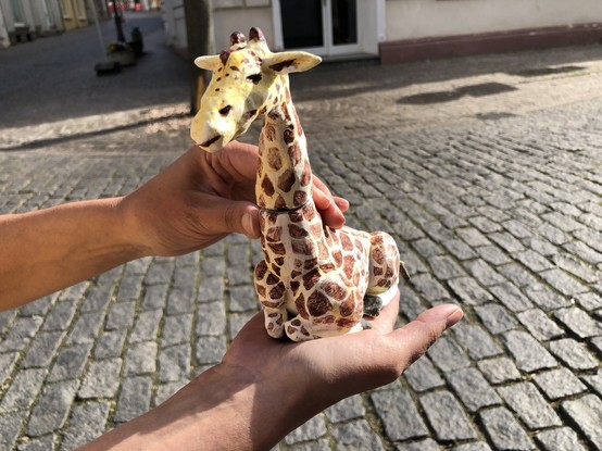 Farbig bemalte und handgefertigte Giraffe (sitzend) aus Ton wird in die Kamera gehalten.