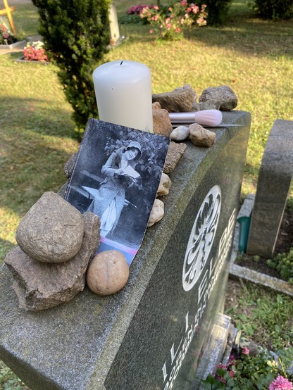 Detailansicht des Grabsteins von Lili Elbe auf dem Trinitatisfriedhof in Dresden-Johannstadt. Auf der Oberseite des Grabsteins sind verschiedene Steine, ein Schwarzweißfoto von Lili Elbe, eine Kerze und ein rosafarbener Make-up-Pinsel abgelegt.