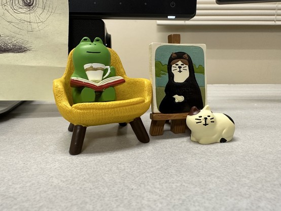 A tiny green frog sits in a yellow chair, with a book and cup of tea. Next to the frog is a tiny cat in front of a portrait of a cat as the Mona Lisa. 