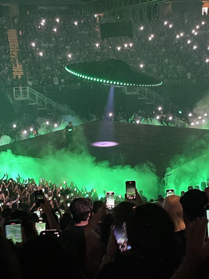 Drake on the center stage, which is fogged up and lit with green light. Over him is a flying saucer, with green running lights and a center beam pointing down to the stage, as if searching for him.