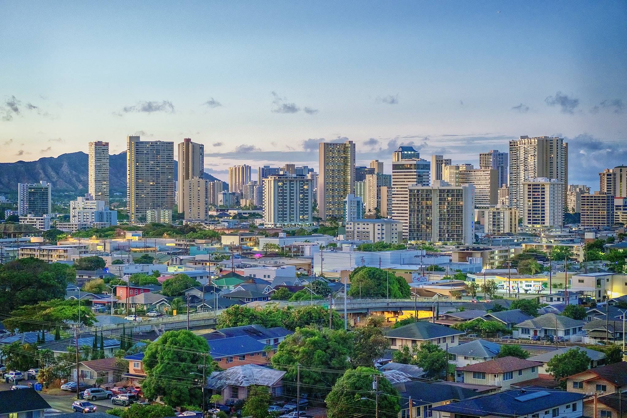 Tall buildings of downtown Honolulu with Diamond Head in the background and residential neighborhoods in the foreground at dawn. Sunlight reflects off the windows of the buildings and the color palette is pastel. 