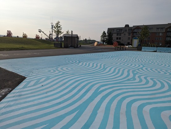 Shows Halifax waterfront. In the foreground is blue and white squiggly lines painted on the ground with condos in the background to the right and small store huts in the background in the center