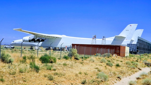 A huge, white airplane is seen parked in rear-quarter view. There are two vertical tails (and though the second is not visible) two long, tapered fuselages. Three huge jet engines hang from the near wing.  