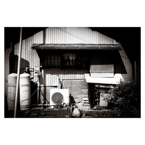 A monochrome image of a humble home with an air conditioner, big gas cylinders, chains, wires, corrugated rusty metal and super high contrast shadows.