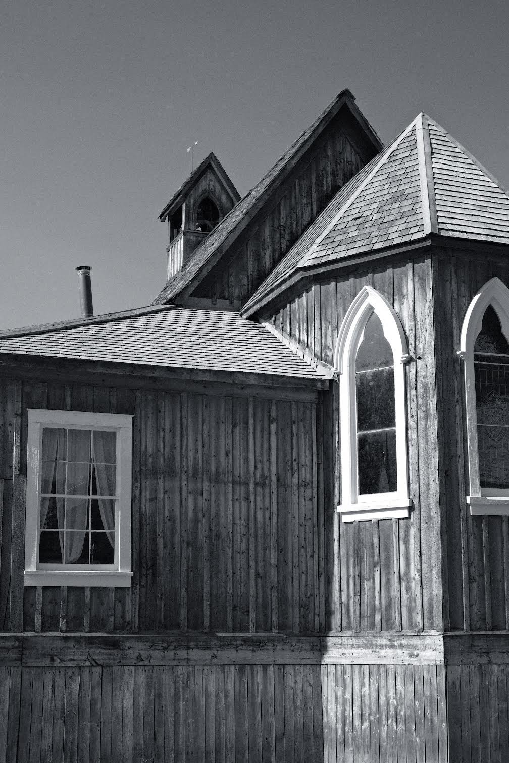 Black and white photo of exterior details of an antique wooden church. Visible are the exterior of the nave, apse and steeple with stained glass windows and sloped rooflines against a cloudless sky.