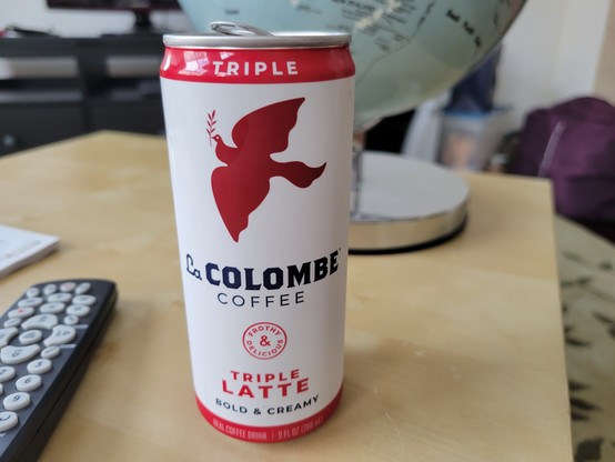9 fluid ounce can of La Colombe Triple Latte Coffee, on a birch veneer IKEA LACK side table. The La Colombe can has a white background with red borders at top & bottom, and a dark red silhouette of a dove of peace in flight while holding an olive branch. Other text on the can: "Triple", "Frothy & Delicious", "Bold & Creamy". Nearby is a TV remote and a Wild & Wolf Light Up Globe.