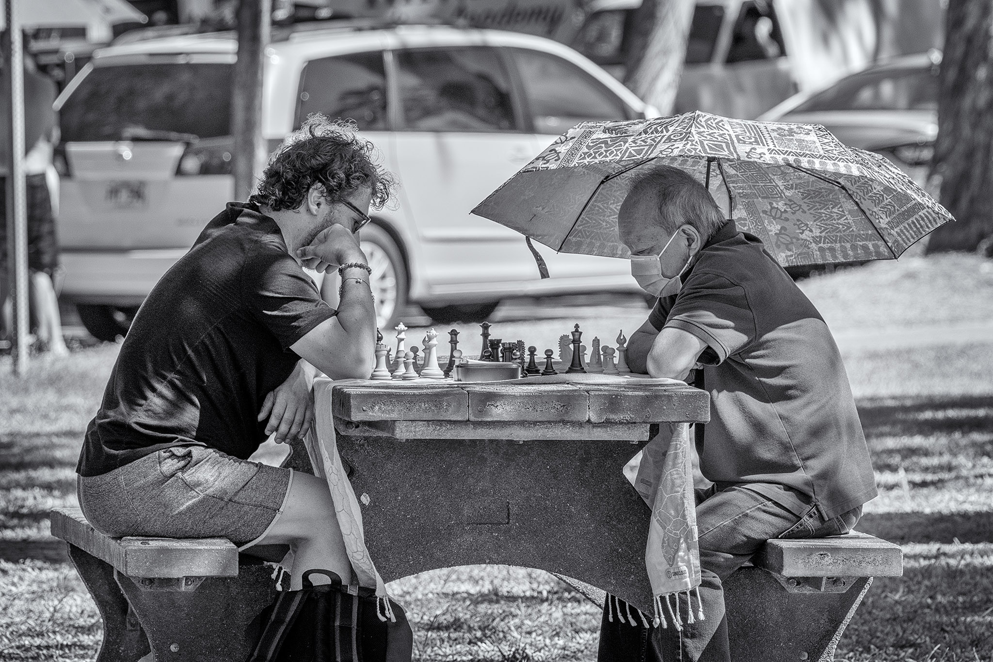 Two men sit at a concrete table in a park near Waikiki with a chess board and pieces between them. One is wearing a mask and holding an umbrella; the other is wearing sunglasses.