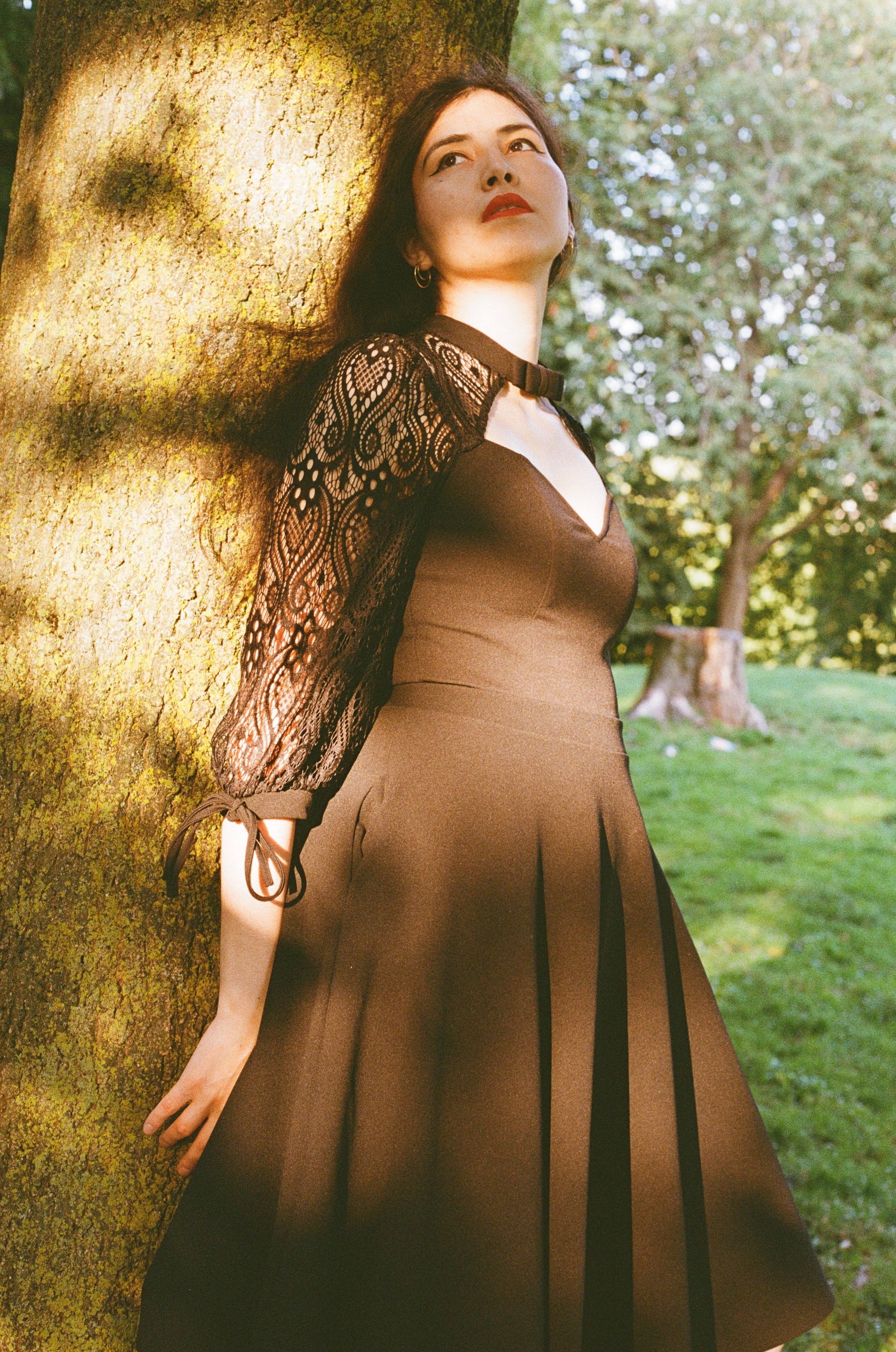 Beautiful woman with long black hair wearing a black dress, leaning against a tree, covered in patches of warm sunlight