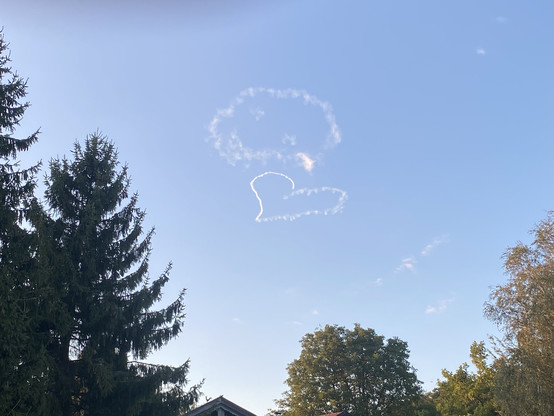 Blick in den blauen Himmel: Ein Kunstflieger hat ein Smiley gezeichnet – das Gesicht ist schon ein wenig verweht, aber noch gut erkennbar. Dazu ein Herz, das noch ganz klare Konturen hat. Am unteren Bildrand sind Baumwipfel zu sehen.