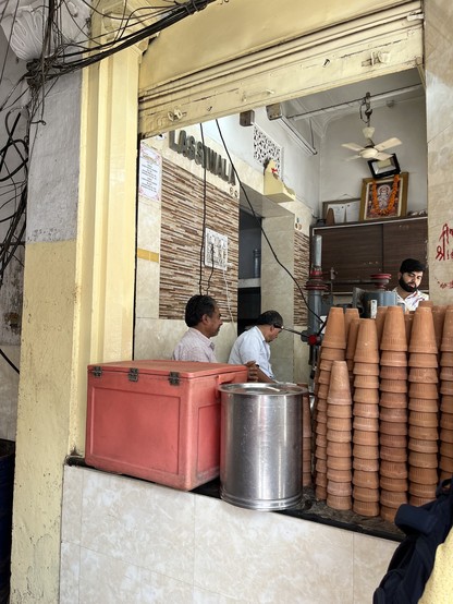 The Real Lassiwala from the side. On the counter of the small stand is a red cooler, a metal basin, and a stack of clay khulads.