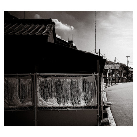 The blazing sun on a hot Nagoya day. There's a chain link fence with tarpaulins in tatters in front of a house with stark shadows.