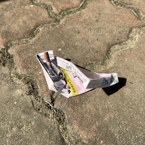 A scrunched up littered cigarette box sitting on sandy pavers in bright sunlight.