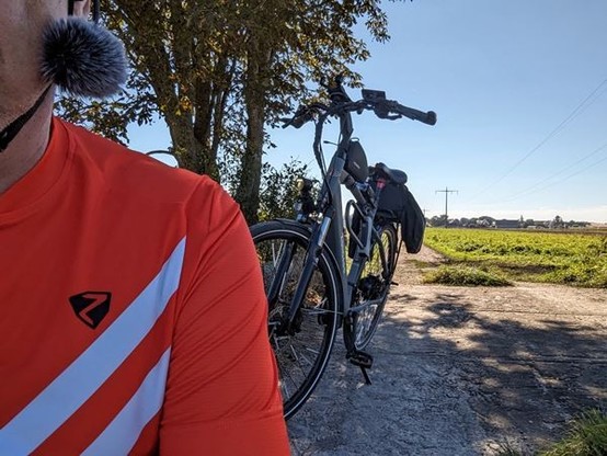 Foto einer Person, die ein orangefarbenes T-Shirt mit einem schwarzen Dreieck trägt und neben einem schwarzen Fahrrad auf einem Feldweg steht. Die Person trägt ein Mikrofon am Kopf. Das Fahrrad ist ein Mountainbike mit schwarzem Rahmen und Lenker. Im Hintergrund ist ein blauer Himmel und grüne Felder zu sehen. Das Foto wurde tagsüber aufgenommen.