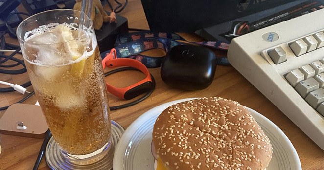 Messy desk with food, soft drink, keyboard, earphones, and whatnot.