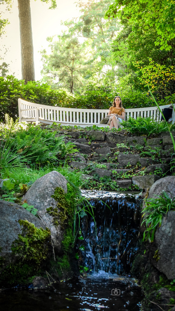 Looking up a small garden waterfall towards a woman wearing a burnt orange T-shirt and a multicolored skirt sitting on a white semicircular wooden garden bench