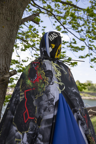 A mannequin draped with banners fixed in place with black cable ties stands ominously beneath a tree in a sunny green landscape.