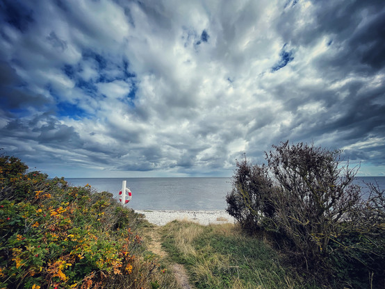 A little footpath leads to a white-cobbled beach on the Baltjc Sea near Grenaa, Denmark. A red-and-white life belt on a post can be seen on the beach, a dramatic clouded sky dominates the background. 