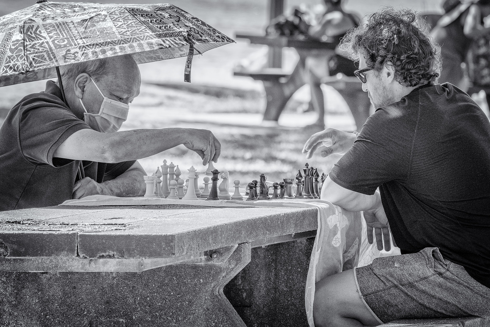 Two men sit at a picnic table in a park in Honolulu playing chess. One is older, wearing a mask and holding an umbrella to block the sun; the other is younger and wears sunglasses.