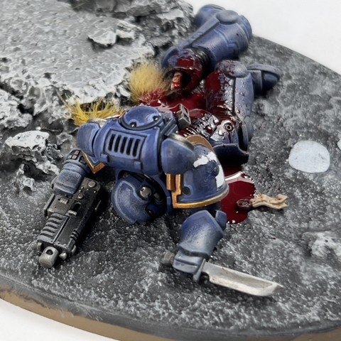 A work in progress shot of a Warhammer 40,000 miniature model. A dead space marine in blue lose dead on the floor, there’s a lot of blood surrounding him.