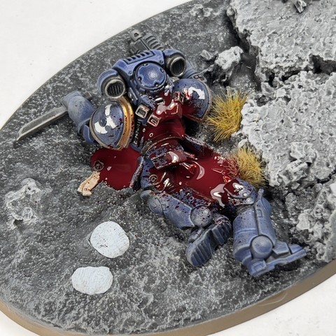 A work in progress shot of a Warhammer 40,000 miniature model. A dead space marine in blue lose dead on the floor, there’s a lot of blood surrounding him.