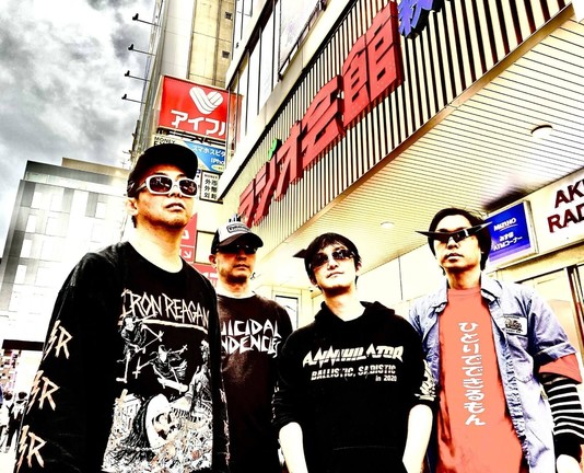 The four men of Satsuriku Robot are standing in front of a building, posing for the camera. They are wearing band shirts or hoodies in mostly dark colours. In addition, they are all wearing sunglasses.