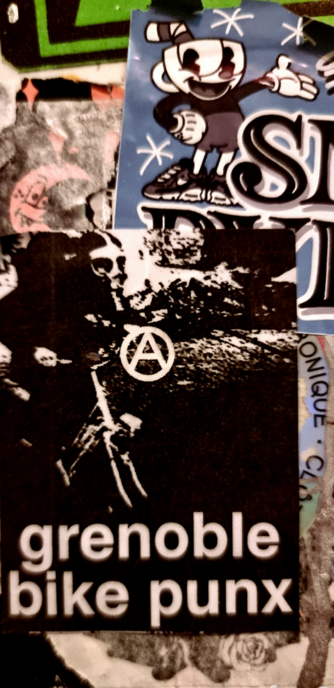 Détail d'un mûr d'autocollants. En bas à gauche, un sticker noir et blanc, avec l'inscription "grenoble bike punx", un A cerclé, symbole anarchiste.