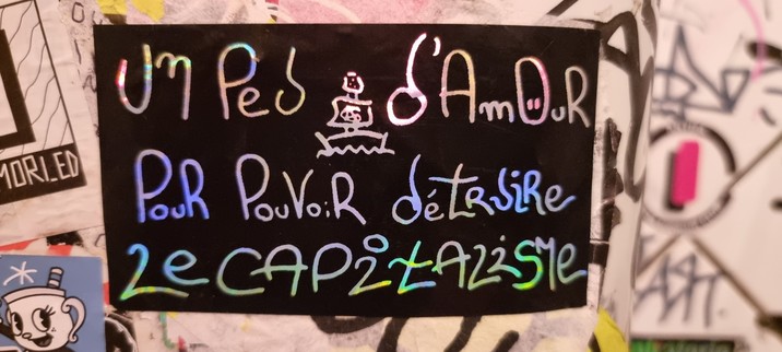 Sticker noir, lettres argentées et reflet arc-en-ciel, pour nous rappeler qu'il faut "Un Peu d'Amour Pour Détruire Le Capitalisme".