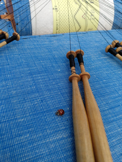 A blue fabric on a lace pillow shows two wooden bobbins wound with black silk thread, and a tiny ladybug right in the way, about a quarter inch from my bobbins.