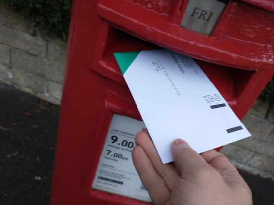 A picture of a red postbox with a label indicating the next collection is Friday. A hand can be seen putting the ballot envelope into the slot of the postbox.