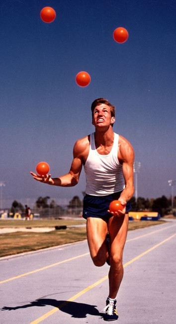 A man in running shorts on a track wearing a white tank top. His expression looks strained and stressed because he's juggling 5 red balls. Juggling white jogging is called joggling. 