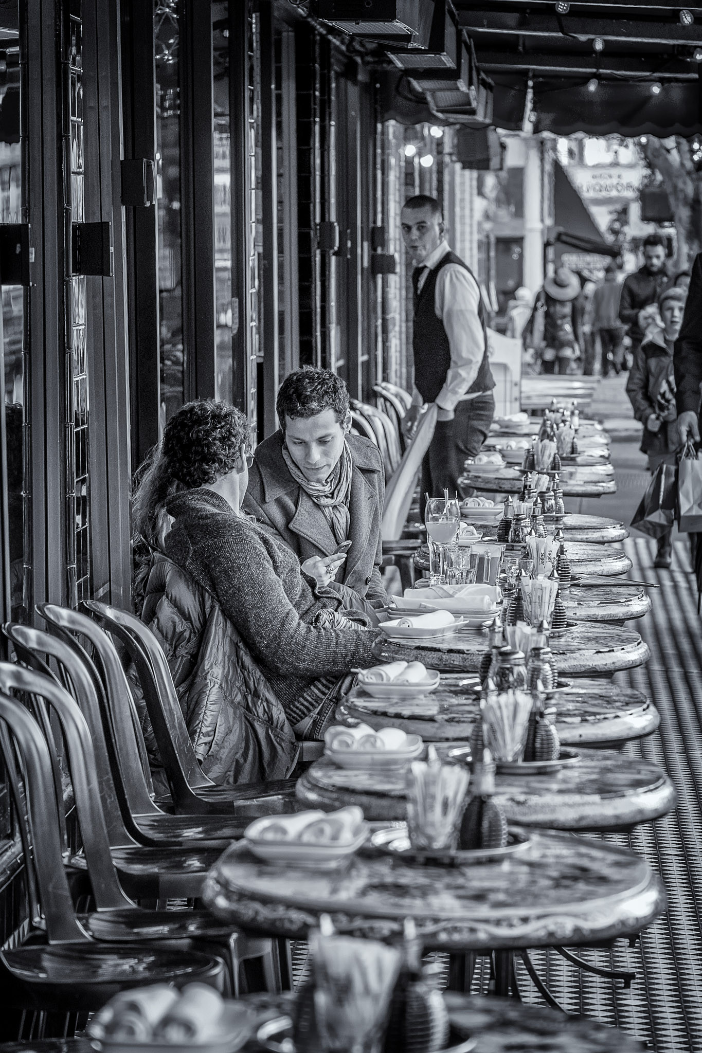 Looking down a row of tables in front of a cafe in the North Beach neighborhood of San Francisco. A couple is sitting in a table in the middle of the row and a waiter looks down the row at the camera.
