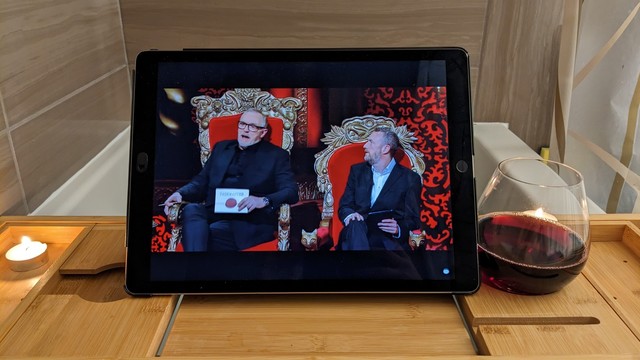 POV photo in a bath. iPad, glass of red wine and two tea light candles on a wooden bath caddy. iPad is playing Taskmaster, shot of Greg Davis and Alex Horne.