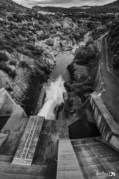 Blick von der Staumauer hinunter ins Flussbett, in das Wasser durch die Staumauer strömt. Im Hintergrund die Landchaft.