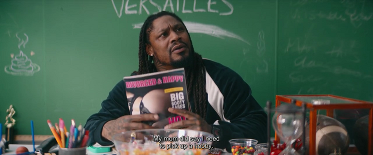 A screen cap from "Bottoms."
Marshawn Lynch, sitting at a teacher's desk at the front of a classroom, is holding a copy of a porno mag called "Divorced & Happy" that identifies that it contains pictures of "big booty babes." He is telling two students who are off-camera "My mom did say I need to pick up a hobby." A poop emoji is drawn on a green chalkboard behind his right shoulder.